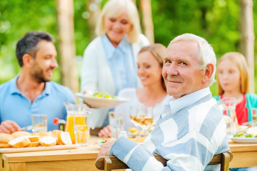 Happy family of five people sitting at the dining table outdoors while senior man looking over shoulder and smiling